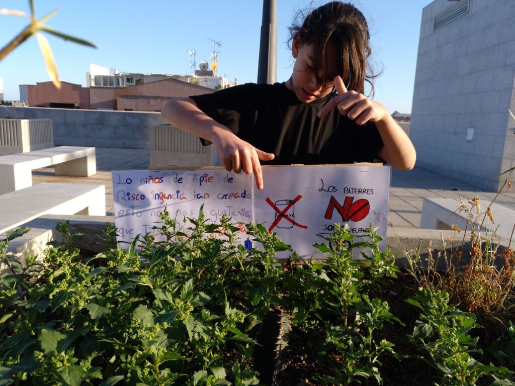 Niña delante de un parterre con plantas y dos carteles hechos a mano por niños y niñas en los que se solicita cuidar las plantas y no tirar basura en el parterre