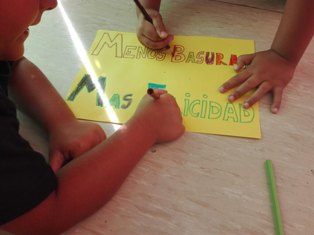 Manos de niños pintando un cartel en el que pone en letras grandes: menos basura, más felicidad